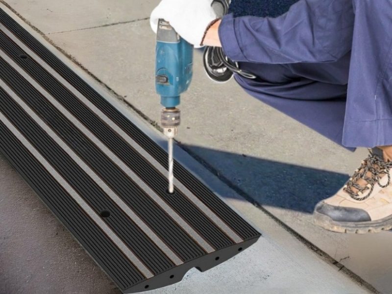 Technician drilling into predrilled holes of a driveway ramp during installation, preparing for secure mounting to a curb to prevent scraping.